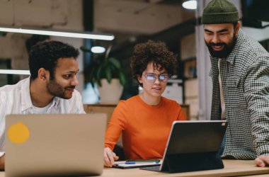 Konsantre olmuş, günlük giysiler içinde laptoplarıyla masada oturmuş, gülümseyen ve aydınlatılmış çalışma alanı projesinde çalışırken ekrana bakan genç meslektaşlar.