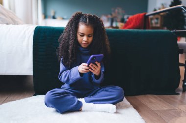 Ciddi Afro-Amerikan kadın çocuk rahat bir yatak odasında dinlenirken bacak bacak bacak üstüne atmış ve çoraplarıyla koltukta oturuyor.