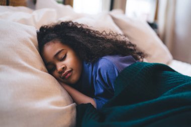 Göz kamaştırıcı kıvırcık saçlı, günlük giysiler içinde battaniyeyle örtülü yastığın üzerinde uzanan parlak yatak odasında gözleri kapalı uyuyan küçük kız.