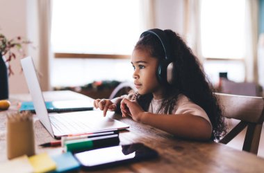 Afro-Amerikan kıvırcık saçlı, günlük kıyafetleri ve kulaklıkları olan, ahşap masada dizüstü bilgisayarla oturup ödevlerini yaparken ekrana bakan kızın yan görüntüsü.