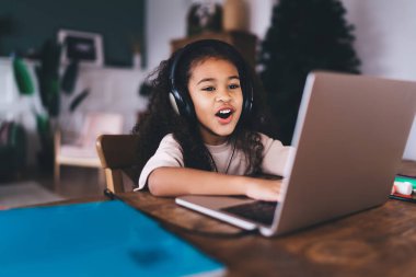 Neşeli Afro-Amerikalı, günlük kıyafetler giymiş, kıvırcık saçlı, dizüstü bilgisayarlı ve ekranda oturup kulaklıkla müzik dinleyen ve ağzıyla şarkı söyleyen kız çocuğu.
