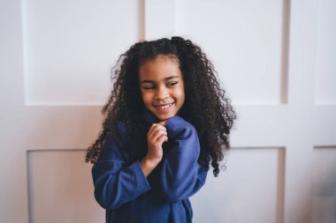 Gülümseyen Afro-Amerikan kıvırcık saçlı kız. Çenesine dokunup mutluluğunu ifade ederken modern apartmanın içinde beyaz bir altyapısı var.