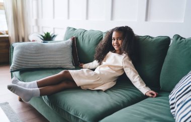 Beyaz çoraplı, tatlı kıvırcık saçlı, rahat bir oturma odasında dinlenirken kameraya bakan neşeli Afro-Amerikan kız.