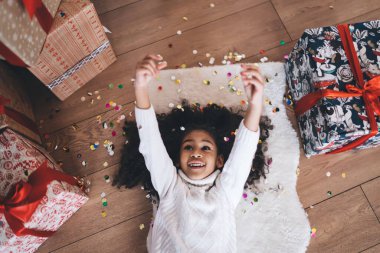 Heyecanlı Afrika kökenli küçük kız sıcak giysiler içinde Noel hediyesi kutularıyla pamuk mindere uzanmış evde tatil boyunca renkli konfetilerle oynarken.