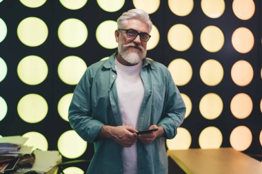 Beyaz sakallı, sıradan kıyafetler ve gözlük takan, elinde akıllı bir telefonla renkli ve modern bir geçmişin önünde duran kendinden emin bir beyaz adam.