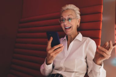 Beyaz gömlekli neşeli beyaz kadın, akıllı telefon kullanıyor, heykel gibi kırmızı bir sandalyeye oturmuş, keyif ve şaşkınlığı ifade ediyor, ofis ortamında.