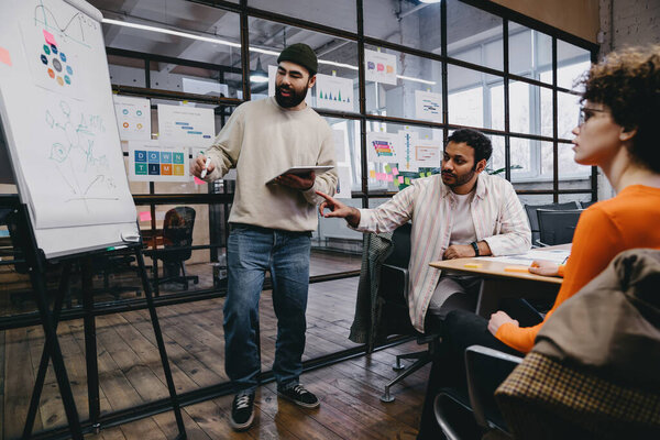 Group of multiracial coworkers in casual clothes sitting at table with papers and discussing details of project while working together on board