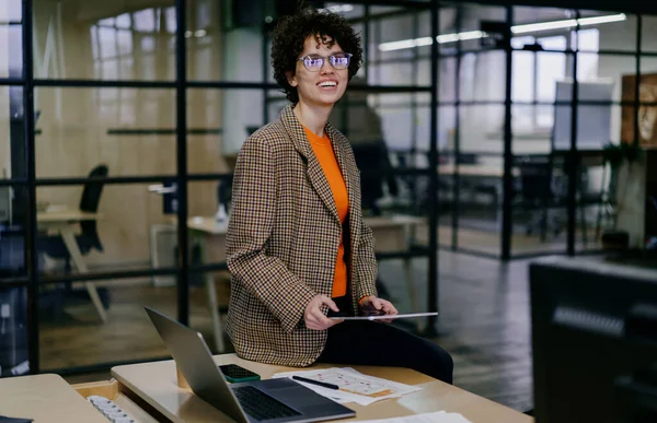 Positive young female entrepreneur in trendy outfit and eyeglasses sitting at table with laptop and tablet and looking at camera while working on project in modern workspace with blurred glass wall