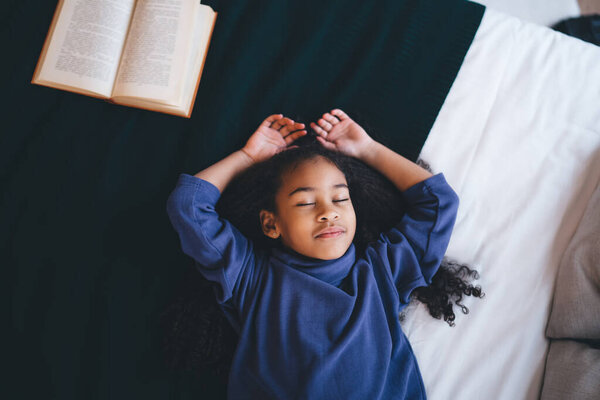 Top view of African American adorable positive girl with curly hair in casual cloth lying with back on bed and closed eyes while thinking over novel