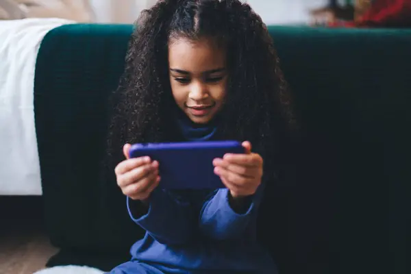 stock image Crop adorable African American female curly hair girl in casual clothes sitting on floor by bed with blankets while playing video games browsing mobile phone
