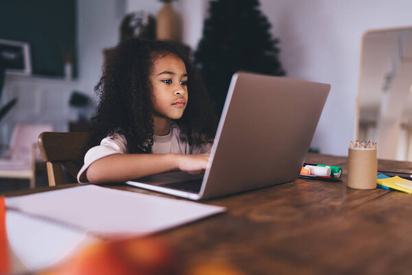 Focused adorable African American female kid in casual clothes sitting at table with laptop and looking at camera while working on remote project at home