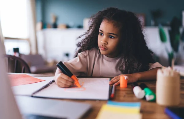 Concentrated African American girl sitting at table thinking and drawing with colored marker on notepad while doing homework over wooden table at home