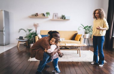 Afrika kökenli Amerikalı bir baba, gündelik kıyafetlerle çömelirken bir yandan da annesi parke zeminin üzerinde dururken sevimli kızına sarılıyor.