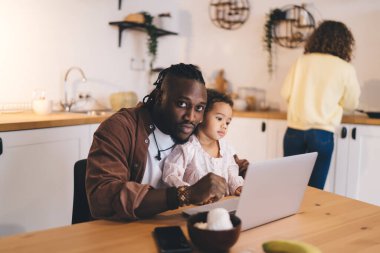 Kendine güvenen Afrikalı, sakallı, saçları kazınmış, sıradan giysileri olan genç bir baba yemek masasında dizüstü bilgisayarla oturmuş kameraya bakıyor.