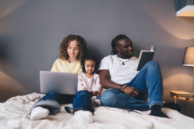 Yatakta dizüstü bilgisayar kullanırken ve rahat bir yatak odasında kitap okurken günlük giysiler içinde konsantre olmuş koca ve eş.