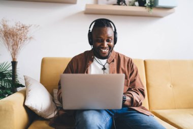 Afro-Amerikalı, kulaklıklı, gülümseyen ve ekrana bakan, koltukta oturup evdeki uzak projeler üzerinde çalışan pozitif Afrikalı erkek serbest yazar.