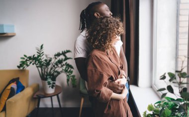 Çok ırklı çiftleri sevmenin yan görünüşü, sıradan giysiler içinde, pencerenin yanında, saksı bitkileriyle dikilip, başka tarafa bakarken, romantik bir şekilde kucaklaşmak ve birlikte oturma odasında vakit geçirmek.