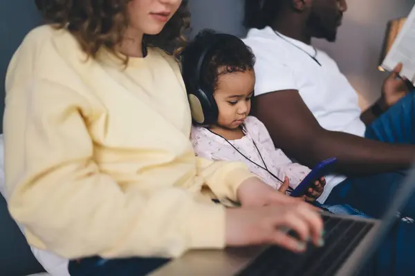Adorable focused African American curly haired girl listening to music over headphones and smartphone while sitting between crop parents on bed in room