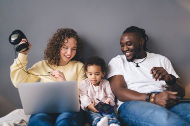 Rahat koltukta oturan, akıllı telefon ve kulaklıklarla bilgisayarı kullanan, aynı odada oynayan ve kaynaşan pozitif, etnik çeşitliliğe sahip çift ve kız.