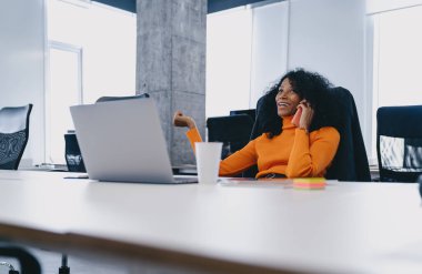 Parlak Afro-Amerikalı iş kadını telefonla gülüyor, modern tasarımla geniş bir ofiste oturuyor, canlı turuncu balıkçı yaka giyiyor, iş ortamında güven ve neşe saçıyor..