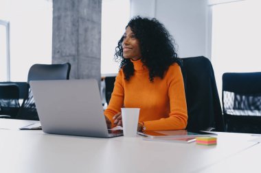 Turuncu kazaklı neşeli bir girişimci, ortak ofis alanında çalışırken hafif bir anın keyfini çıkararak, girişimci çalışma ortamlarının pozitif kültürünü yansıtıyor..