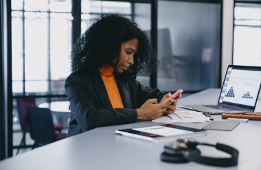Siyahi iş kadını, kıvırcık saçlı, iş yerinde akıllı telefonunu kullanıyor, ofis masasında dizüstü bilgisayarı ve belgeleriyle, güneşli, modern ofis pencerelerinin önünde..