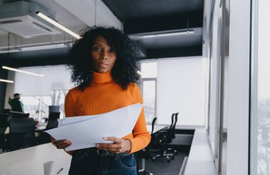 Cesur turuncu kazaklı bir analist, parlak bir ofiste finansal raporları inceliyor. Bu, yoğun bir şirket ortamında odaklanmış iş değerlendirmesi örneğini veriyor..