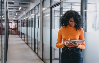 Genç siyahi iş kadını modern ofis koridorunda tablet kullanıyor, odaklanmış ve ciddi. Günlük iş kıyafetleri giyerek, modern sektörde profesyonelliği, teknolojiyi ve hareketliliği somutlaştırıyor..
