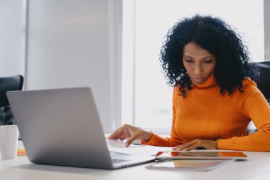 İşletme analizlerini tablet üzerinde gözden geçiren konsantre Afrikalı Amerikalı girişimci, birlikte çalışma, laptop açık ve kağıt, turuncu süveter iş ayarlarına biraz canlılık katıyor.