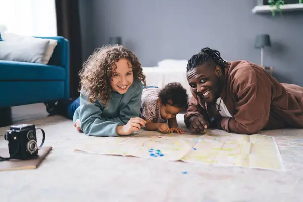 Happy multiethnic family with toddler kid lying on carpet near photo camera and pinning on map while spending time at living room in apartment against blurred background