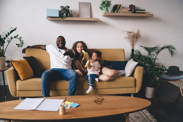 Cheerful multiracial family in casual clothes sitting on sofa and eating banana while making hand gestures and spending time together in cozy living room with wooden table