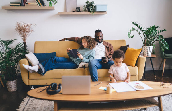 Cheerful multiracial couple and daughter in casual clothes sitting together on sofa and reading book while browsing smartphone and spending time together at home with cute little girl