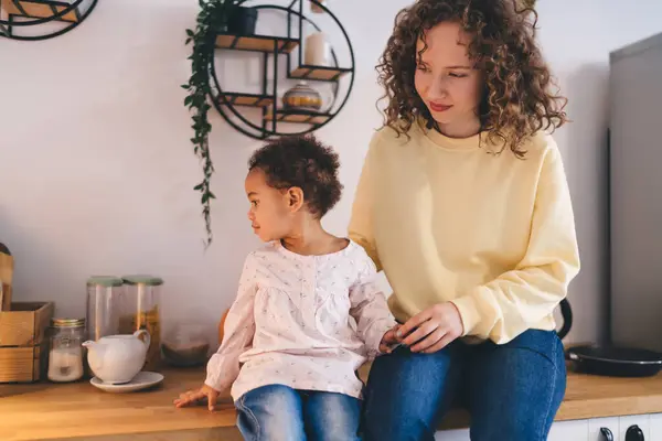 Calm mother with cute little girl in curly hair and casual clothes with jeans sitting on wooden kitchen table with teacup while spending time together at home
