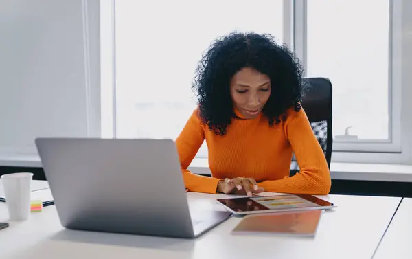 stock image Engrossed African American cryptocurrency trader, analyzing live digital currency charts on tablet, with laptop beside in coworking, orange sweater symbolizing the energetic pulse of crypto market.