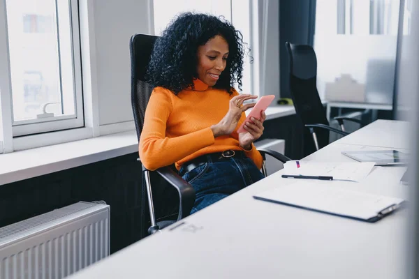 Smiling Black businesswoman in her 30s, enjoying a light moment with her smartphone in a sunny, modern office, reflecting a relaxed work environment in autumn.