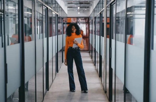 Black businesswoman in vibrant orange sweater and black jeans with a digital tablet in an office hallway, exemplifying modern business mobility and style.