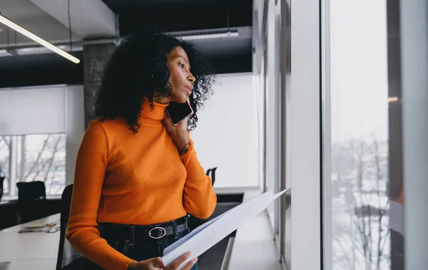 A travel consultant in an eye-catching orange sweater carefully reviews vacation policies on a call, planning eco-friendly itineraries in a bright, modern office.