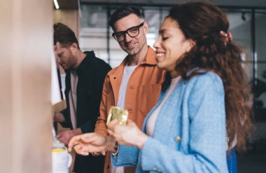 Bir grup çok ırklı iş arkadaşı şık giyinmiş, dinlenme yerinde kahve yapıyor ve iş arasında gülümsüyor.