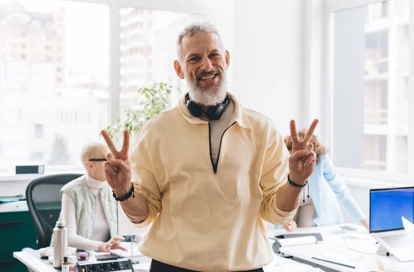 Cheerful bearded male employee in casual clothes with headphones on neck showing peace gesture while smiling and standing against coworkers in light office