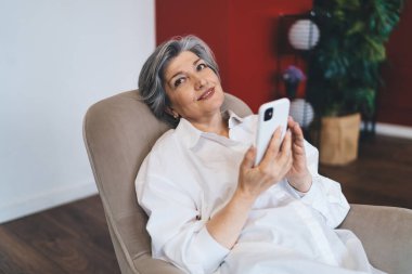 Pozitif orta yaşlı, gri saçlı, sıradan elbiseli, gülümseyen ve telefon kullanan bir kadın. Oturma odasındaki kanepede oturuyor ve başka tarafa bakıyor.