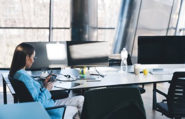Gözlüklü, resmi giysileri olan, bilgisayar masaüstleriyle masada oturan ve ofisteki projede çalışırken akıllı telefonları karıştıran yoğun saçlı kadın ofis çalışanının yan görüntüsü.