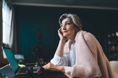 Düşünceli, orta yaşlı, serbest çalışan, sıradan elbiseli, gülümseyen ve yüzünü çeviren bir kadın.