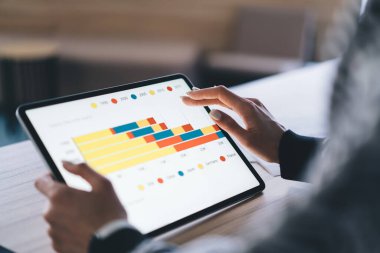 Ahşap masada oturmuş, parlak renkli bar çizelgesiyle tableti karıştırırken ve modern çalışma alanında proje üzerinde çalışırken gündelik giysiler içinde anonim girişimcinin yukarısından