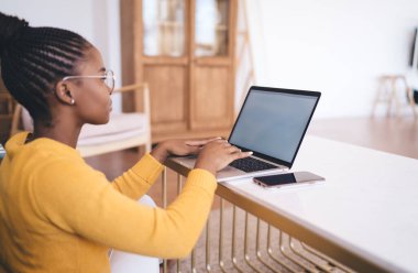 Saçları örgülü, akıllı telefonuyla masada oturan ve evde çalışırken bilgisayar ekranına bakan Afrikalı Amerikalı serbest kadın.