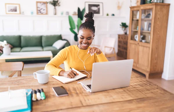 Cheerful young black female student looking down and writing notes with pen in notebook while sitting at table with laptop smartphone and listening to lesson on wired earphones at home