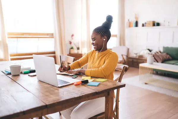 Side view of smiling African American female freelancer with braided hair sitting at table with laptop markers and looking down while writing notes and listening in earphones in daylight