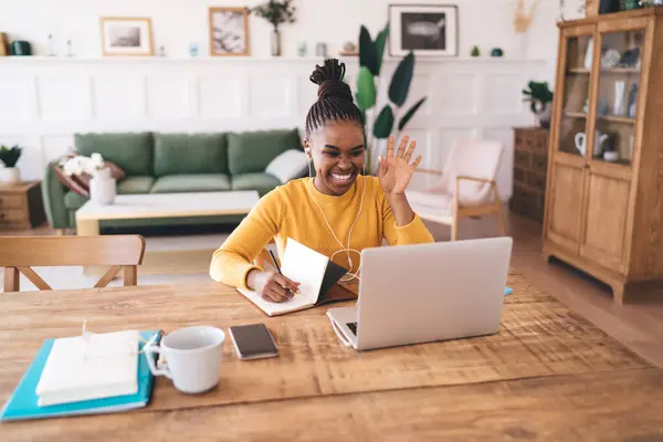 Smiling young black female in braided hair attending video call and saying hello while looking at screen of laptop on table with smartphone and taking notes at home