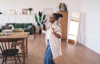 Moda kıyafetleri içinde aktif siyahi bir kadının evde kulaklıklı cep telefonuyla müzik dinlerken son derece mutlu olması.