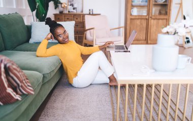 Yan görünüş: Genç Afrikalı Amerikalı kadın serbest çalışan günlük kıyafetler ve gözlüklerle yerde oturuyor ve evden proje üzerinde çalışıyor.