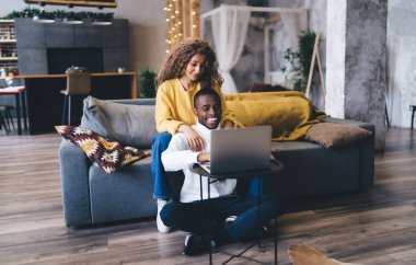 Beyaz balıkçı yaka yaka giyen siyah bir adam dizüstü bilgisayar kullanarak yerde oturuyor, gülüyor, 20 'li yaşlarının ortasında kanepesinde oturan İspanyol bir kadın, kollarını omzuna atmış, rahat bir anın tadını çıkarıyor.